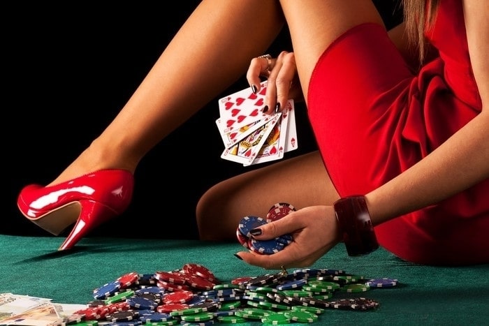 A close-up of a woman’s hands holding playing cards and poker chips on a green xxx roulette table, wearing a red dress and high heels.